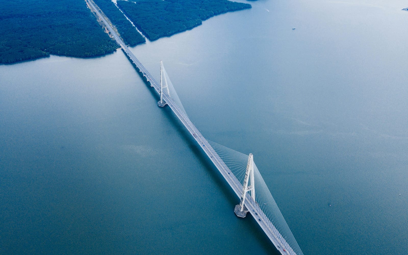 Aerial view of a cable-stayed highway bridge spanning a wide expanse of teal water between forested banks.