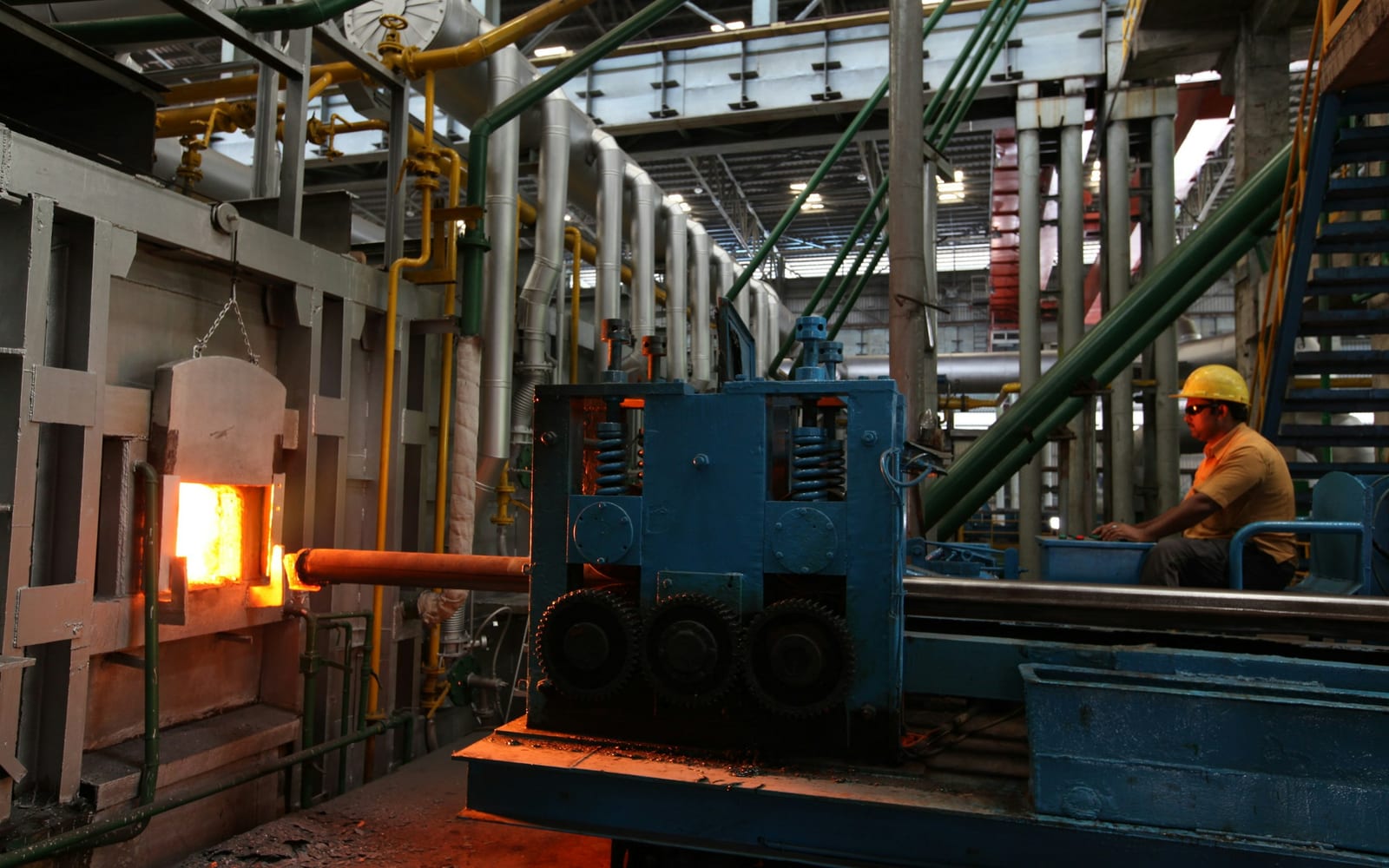 Steel mill interior with a glowing orange furnace, blue industrial machinery, and a worker in a yellow hard hat.